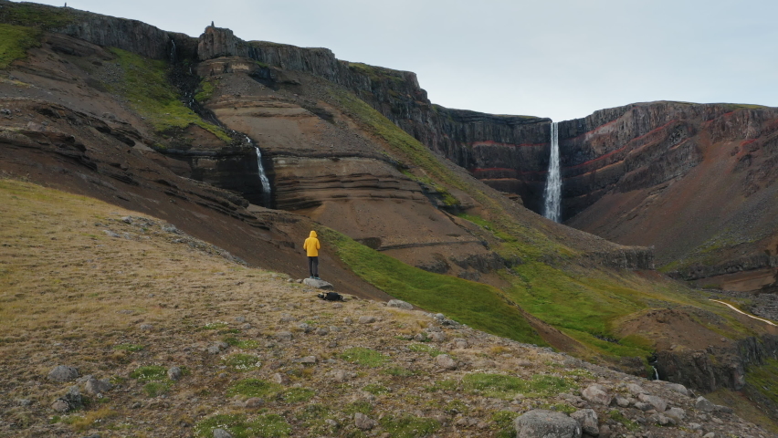 Hengifoss Waterfall, Iceland. Aerial fly above man tourist in yellow jacket on hill enjoying canyon and mountain river