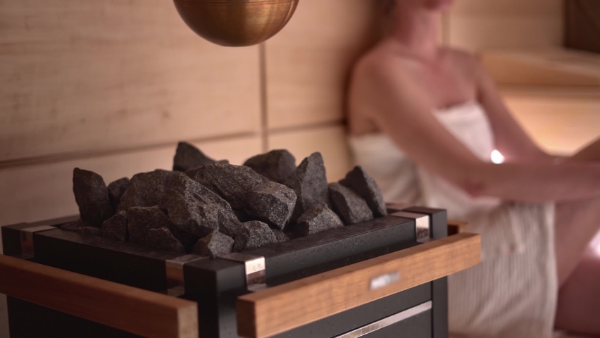 Woman relaxing and sweating in hot sauna. Real, authentic sauna moment. Focus on sauna stove and stone. Royalty-Free Stock Footage #1065288466