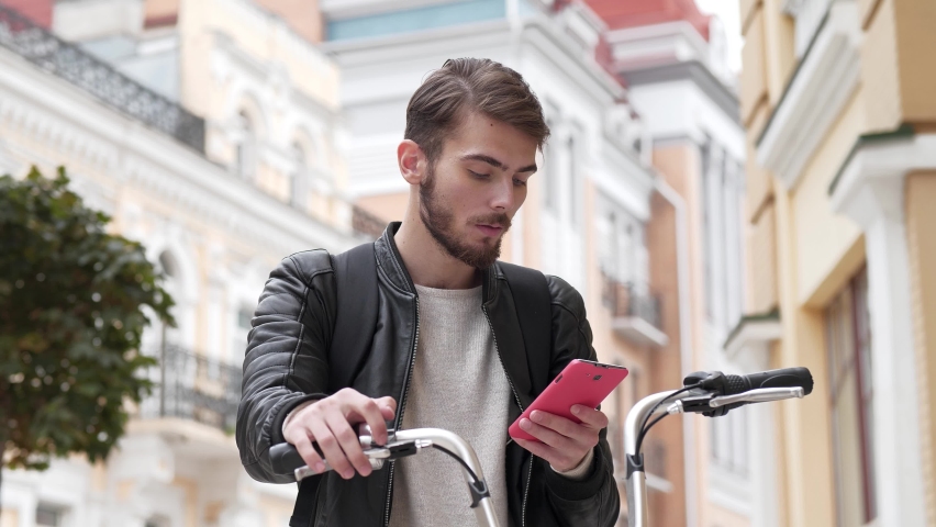 Slow Motion Young man stands on the street with bicycle and reads a message on his smartphone, then he celebrates victory and shows violent emotions