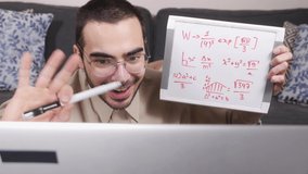 Holding a white board, a teacher is teaching via Internet in his livingroom using a laptop computer.