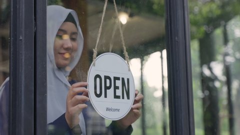 Young Asian Muslim woman wearing hijab, turning sign to open the coffee shop or restaurant. Cheerful female Muslim business owner changing sign from closed to open.