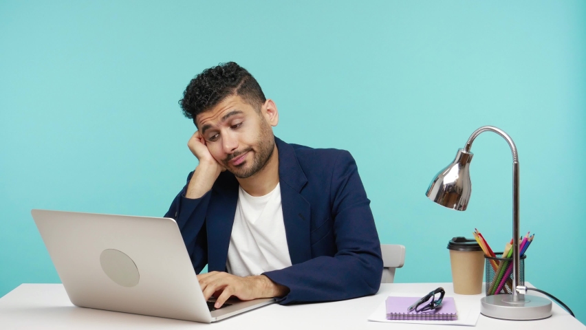 Bored indifferent office worker in elegant suit sitting on his workplace looking at laptop display with disinterest, procrastination. Indoor studio shot isolated on blue background | Shutterstock HD Video #1065305407