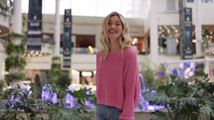 Attractive blonde girl dancing, look at the camera and smiling. Young woman jumping for joy with colorful bags in hands after successful shopping at store. Happy female having fun in shopping mall.
