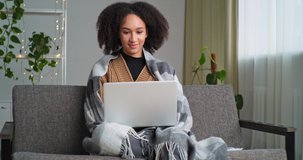 Afro American girl student sits at home living room on couch, covered with blanket holds laptop answers video call speaks remote on online conference waves greeting to webcam, quarantine communication