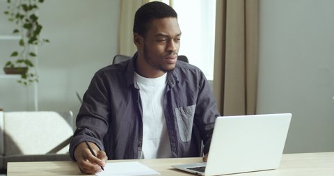 Young guy student pensive afro american business man freelancer sits at table in front of laptop works studies writes with pen on white paper in document plans analyzes does paper work in home office 