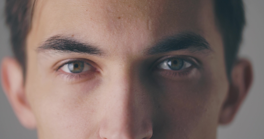 Close-up view of a calm young man is opening his eyes and looking to the camera at grey background wall in studio