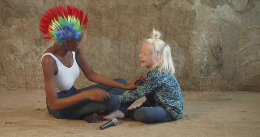 African American woman in white bodysuit and jeans in bright multicolored punk wig and white blond boy singing karaoke with microphone and fooling around. Young mother and foster child love each other