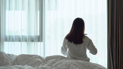 Back view of young Asian Woman sitting on bed near the big white window while stretching after waking up with sunrise in the morning. 