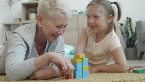 Medium shot of attractive elderly woman and her handicapped granddaughter lying on carpet in living room and playing with colorful tinker toys