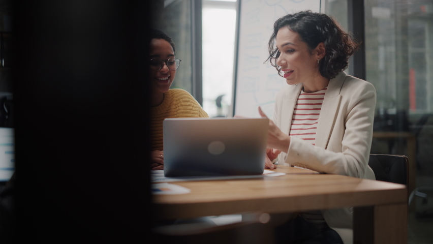 Two Diverse Multiethnic Female Have a Discussion in Meeting Room Behind Glass Walls in an Agency. Creative Director and Project Manager Compare Business Results on Laptop and App Designs in an Office. | Shutterstock HD Video #1065327916