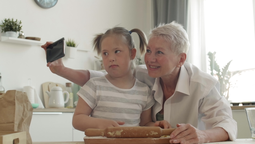 Lockdown of blond-haired old Caucasian woman making selfie with her granddaughter with down syndrome while baking pie