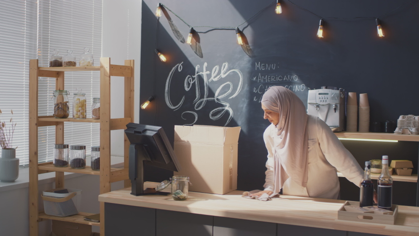 Medium shot of female muslim coffee seller at workplace cleaning cash desk getting ready for opening cafe