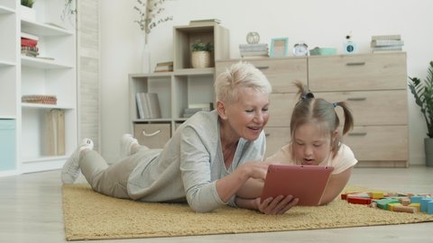 Wide shot of blond-haired Caucasian aged woman and handicapped girl lying on carpet in living room and using digital tablet together