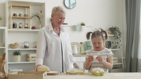 Medium shot of cute teenage girl with down syndrome standing at kitchen table and putting eggs into bowl of flour while her grandmother watching