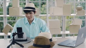 Smiling Asian man influencer reviewing fedora hat and selling a product online at home during COVID-19 pandemic.