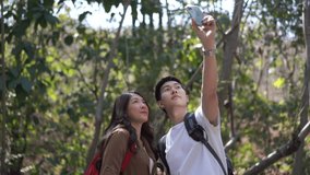 Happy young Asian couple hiking in forest together with using smartphone taking selfie. Smiling man and woman relax and enjoy with outdoor lifestyle. Technology and summer holiday vacation concept 