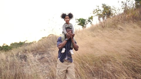 Happy mixed race family on holiday vacation. Smiling father and little daughter carrying and hiking together on meadow hill in summer. Parent with cute child girl relax and enjoy outdoors lifestyle.