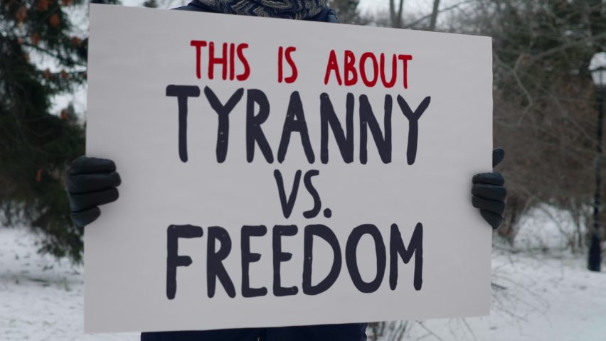 A male protester in winter clothing holding a board with writing This is about Tyranny vs Freedom. This message supposed to show the real meaning behind the popular political parties. Single picket.