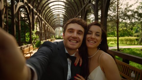 Cheerful man and woman saying hi on phone camera outdoors. Pov shot of happy wedding couple having video call on mobile phone in park. Smiling bride and groom showing rings on camera under arch.