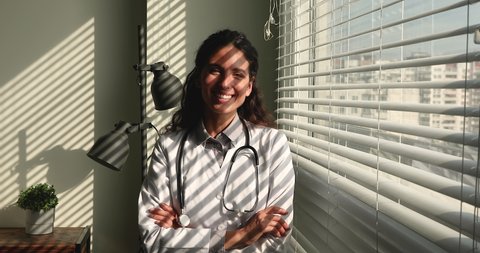 Female general practitioner in white coat with stethoscope on neck smile look at camera, sunlight through window blind. Portrait of confident cardiologist with arms crossed start workday at hospital