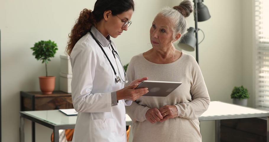 During visit young female doctor in white coat showing to older female patient health test result, explain prescription using medical app on digital wireless tablet. Elder care, modern tech concept Royalty-Free Stock Footage #1065349579