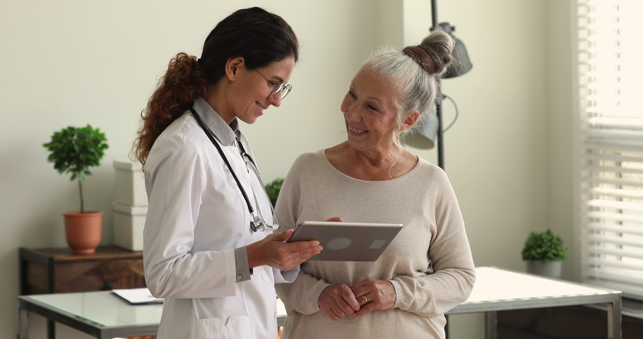 During visit young female doctor in white coat showing to older female patient health test result, explain prescription using medical app on digital wireless tablet. Elder care, modern tech concept | Shutterstock HD Video #1065349579