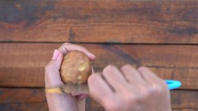 Closeup top view 4k video of woman peeling fresh young spring potato isolated on brown wooden background. Woman scrapes thin skin of vegetable
