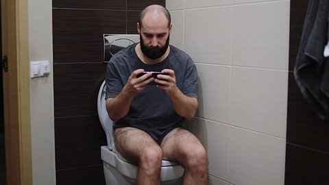 Man Sitting On Toilet Use Cell Smart Phone Young Latin Guy Playing Game In Restroom