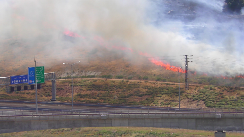 Mount carmel is burning and there is a lot of smoke from the strong wind, cars driving below in nearby highway and road signs