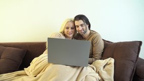 Happy married couple relaxing on comfortable sofa, watching funny video on laptop. Excited smiling spouses laughing on comedian movie