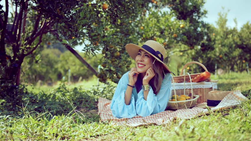 Young asian attractive female wearing casual dress sit relax picnic in orange fresh garden farm ,Girl on a picnic with sandwiches, fruits and drinks. Farm girl with lunch.