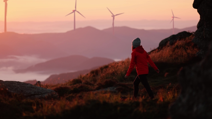 Young woman in orange jacket running up on top of mountain summit at sunset, raises arms into air, happy and drunk on life and happiness. Girl stand against landscape with wind turbines on background Royalty-Free Stock Footage #1065360532