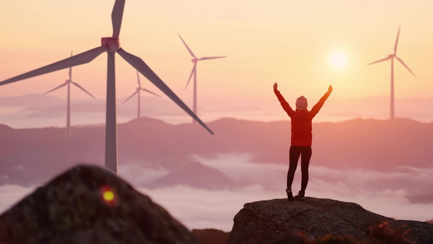 Young woman in orange jacket running up on top of mountain summit at sunset, raises arms into air, happy and drunk on life and happiness. Girl stand against landscape with wind turbines on background | Shutterstock HD Video #1065360532