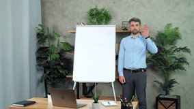 Confident coach trainer greets records online business webinar master class course in office. Man teacher tutor stands near whiteboard listens student question tells teaches remote video call web chat