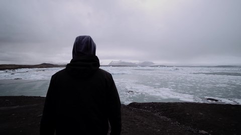 Lonely man walking towards frozen melting lake. Harsh winter landscape, sea covered in ice. Climate change, global warming. Isolated traveler in Iceland.