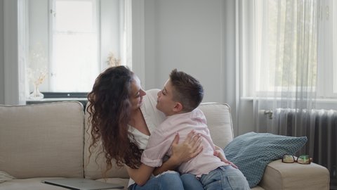 Young Mother And Son On The Couch. Mom Hugs The Boy. They Have Fun. Beautiful Interior In Light Colors. Pretty Young Woman With Long Curly Hair. Woman Kisses The Boy On The Nose.