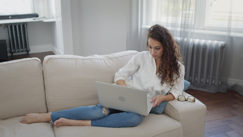 Young Mother And Son. Young Woman Lying On Sofa With Laptop. Beautiful Interior In Light Colors. Young Beautiful Woman With Long Curly Hair. Boy Runs To His Mother And Hugs Her.