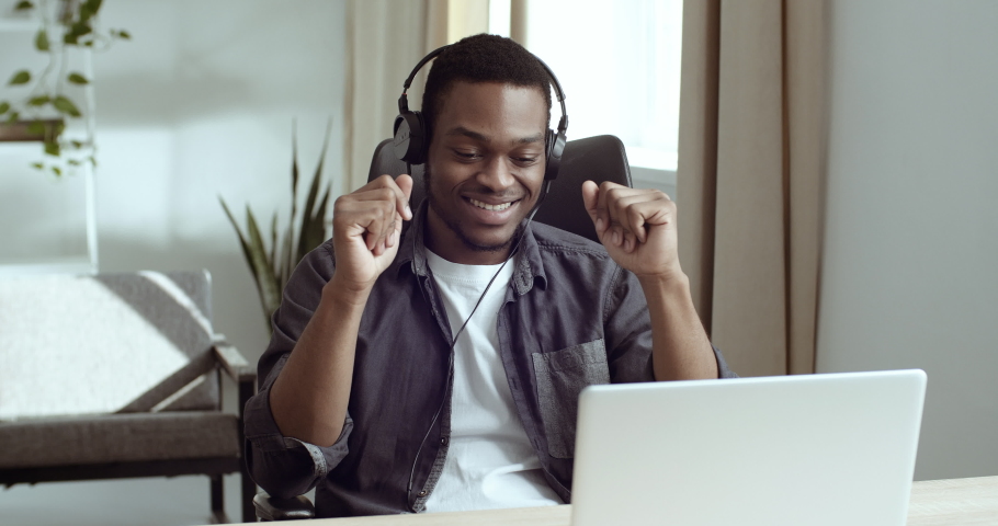 African business man sits at table at his workplace in front of laptop wears headphones listening to music pretends to play drums moves his hands like musician enjoys song resting on break at home | Shutterstock HD Video #1065373942