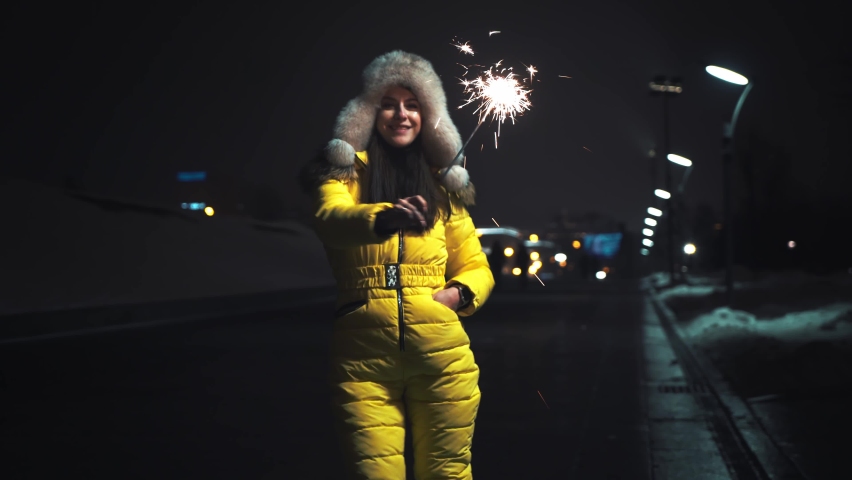 Young cute girl in a winter fur hat and yellow jacket in a festive mood. Sparkling burning sparklers in hand with flying sparks. Dancing and having fun. Holidays concept. Moving camera