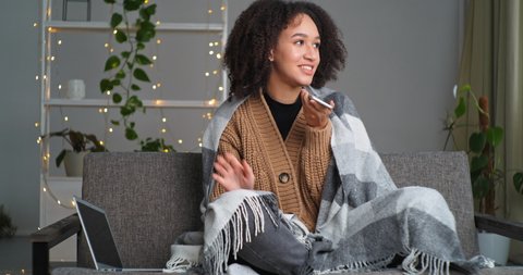Young afro american girl teenager with curly hair sits on sofa in cozy home interior covered with woolen blanket speaks on phone communicates with friends family smiling enjoys dialogue laughs