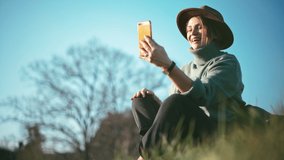 A beautiful middle-aged woman in a hat is taking a video call on her smartphone while sitting on the grass at the park on a sunny day.