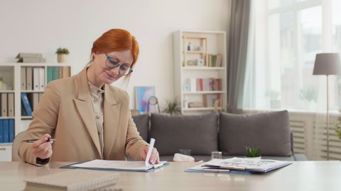 Medium close-up of red-headed small Caucasian female psychologist wearing beige blazer and glasses, sitting by desk in her office, filling in papers with pen, smiling