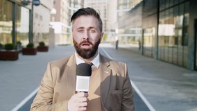 Waist up shot of Caucasian male TV reporter looking at camera and talking into microphone while standing outdoors on city street