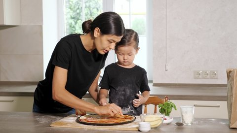 Mother shows how to smudge delicious ketchup on homemade thin base for pizza with tablespoon to little girl in black shirt stained with flour