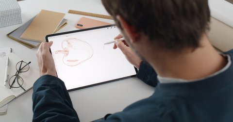Lviv, Ukraine - May 28, 2020: Over shoulder view of male graphic designer creating illustration on tablet.Talanted illustrator using digital pad while sitting at workplace and making sketch.