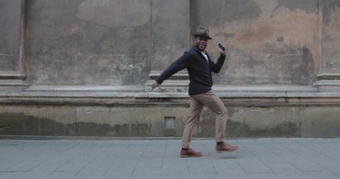 Handsomemmale person dancing and rejoicing while using smartphone at city street. Millennial afro american man in hat walking and smiling while moving in rhytm. Concept of emotions.