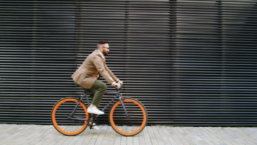 Side view medium shot of Caucasian businessman in smart casual outfit riding bicycle along urban wall on city street while going to office in the morning Royalty-Free Stock Footage #1065402631
