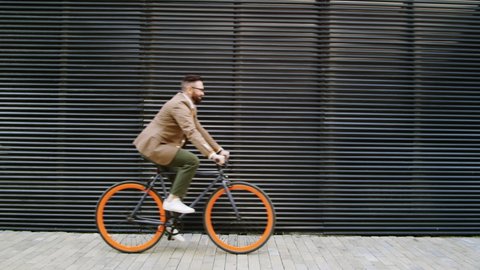 Side view medium shot of Caucasian businessman in smart casual outfit riding bicycle along urban wall on city street while going to office in the morning
