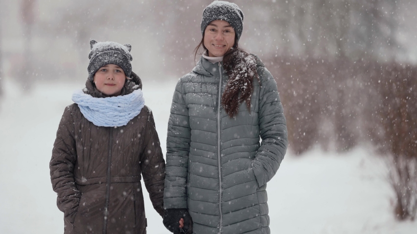 A little girl in a hat and jacket stands with her mother by the hand in the open air on a winter day. They smile. Snow falls, snowy weather in the park.