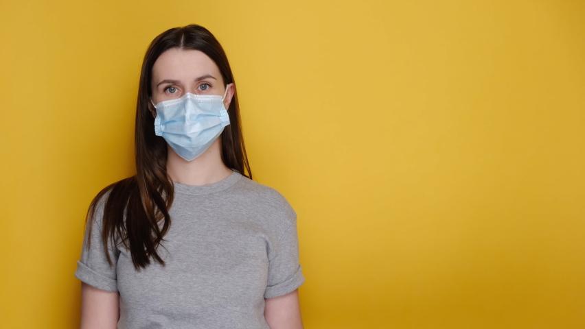 Young female in medical mask and latex gloves showing aside with arrow with thumbs up like gesture, dressed in grey t-shirt, isolated on yellow wall with copy space for advertisement. Covid 19 concept